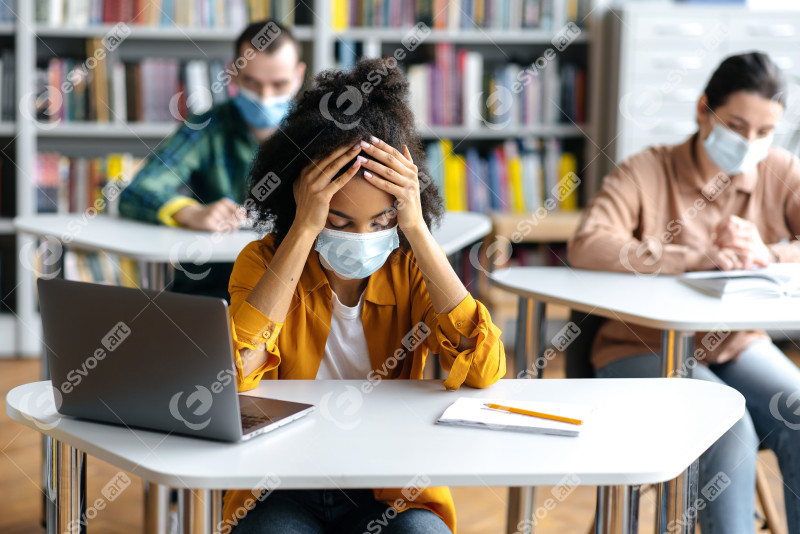 Education during pandemic. Frustrated, overworked african american student girl wearing protective medical mask sits at a table in the library, preparing for exams, tired, classmates sit at background