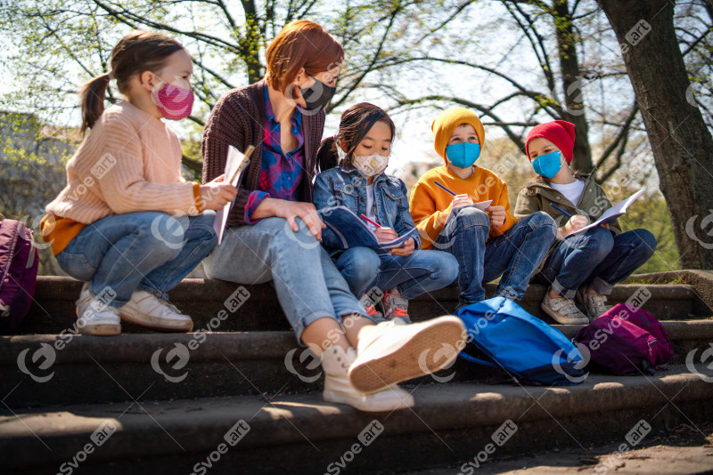 Teacher with small children sitting outdoors in city park, learning group education and coronavirus concept.