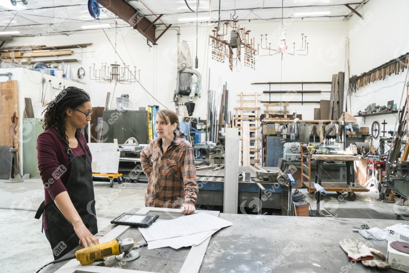 Two women standing at workbench in metal workshop.